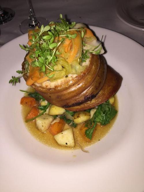 A gourmet dish featuring a layered meat preparation topped with vegetables and microgreens, served on a bed of sauce.