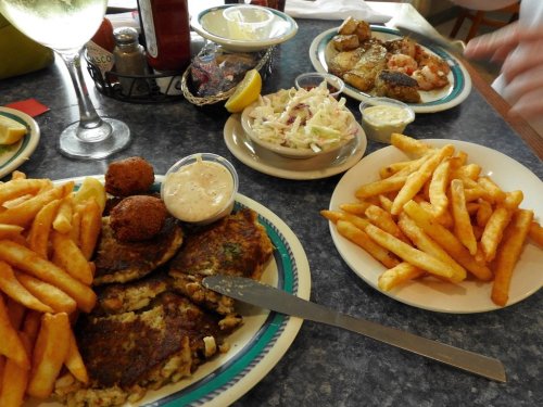 A table with two plates of food: one with fries and seafood cakes, the other with coleslaw and shrimp. Drinks in the background.