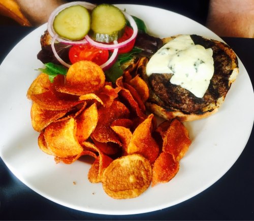 A plate with a burger topped with sauce, lettuce, tomatoes, onions, pickles, and a side of crispy potato chips.