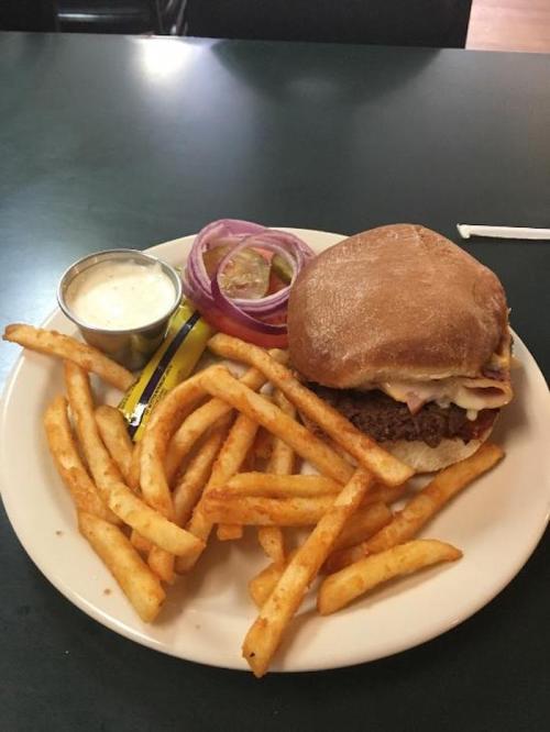A plate with a burger topped with cheese and ham, served with fries, pickles, and a small cup of dipping sauce.