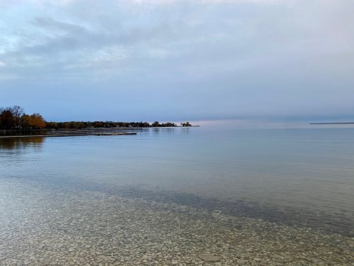 Calm lake scene with clear water, a rocky shore, and a cloudy sky reflecting soft colors at dusk.