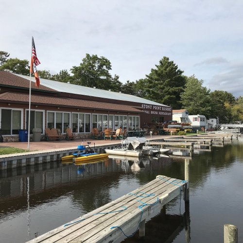 A lakeside resort with a restaurant, docks, and boats, surrounded by trees under a cloudy sky. American flag visible.
