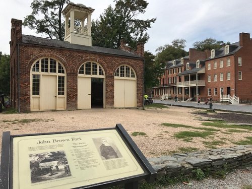 Historic brick building with a bell tower, sign about John Brown Fort, and nearby red brick structures in a park setting.