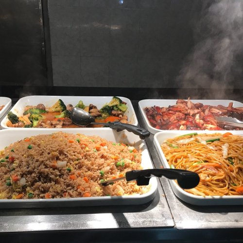 A buffet display featuring fried rice, stir-fried vegetables, chicken, and noodles, with steam rising from the dishes.
