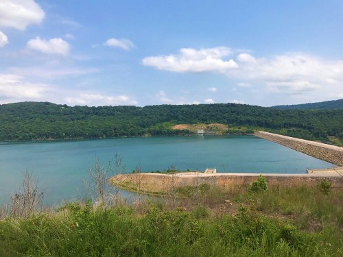 A serene lake surrounded by green hills under a partly cloudy sky, with a stone pier extending into the water.
