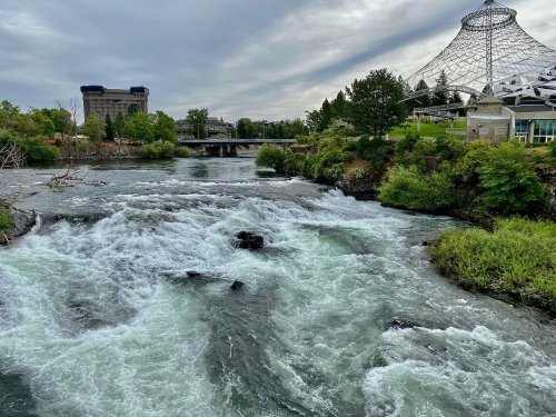A river with rapids flows through a green landscape, with a modern structure and cloudy sky in the background.