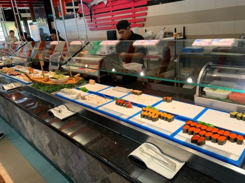 A sushi bar display featuring various sushi rolls and ingredients, with chefs preparing food in the background.