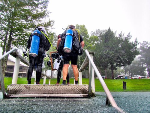 Two divers in wetsuits ascend a metal staircase, carrying scuba gear, as rain pours down around them.