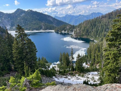 A serene mountain lake surrounded by evergreen trees and snow-capped peaks under a partly cloudy sky.