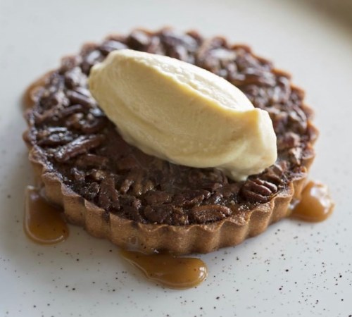 A pecan tart topped with a scoop of vanilla ice cream, drizzled with caramel sauce on a white plate.