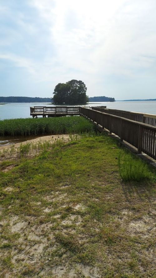 A wooden pier extends over a calm lake, surrounded by grass and trees under a cloudy sky.