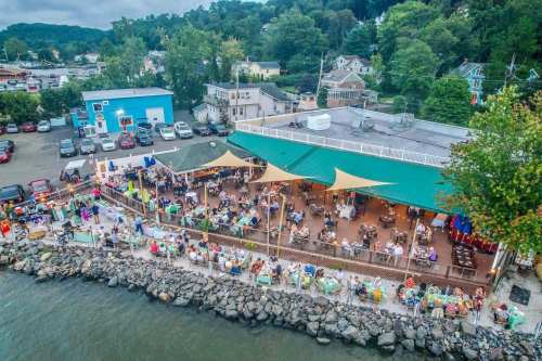 Aerial view of a waterfront restaurant with outdoor seating, surrounded by trees and parked cars.