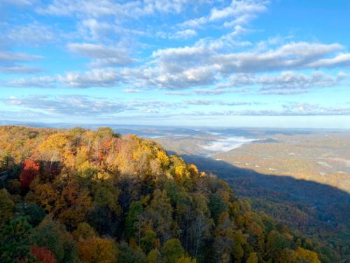 A scenic view of colorful autumn trees on a hillside, with rolling mountains and a cloudy blue sky in the background.