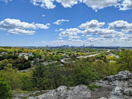 A panoramic view of a city skyline surrounded by greenery and a winding road under a blue sky with fluffy clouds.