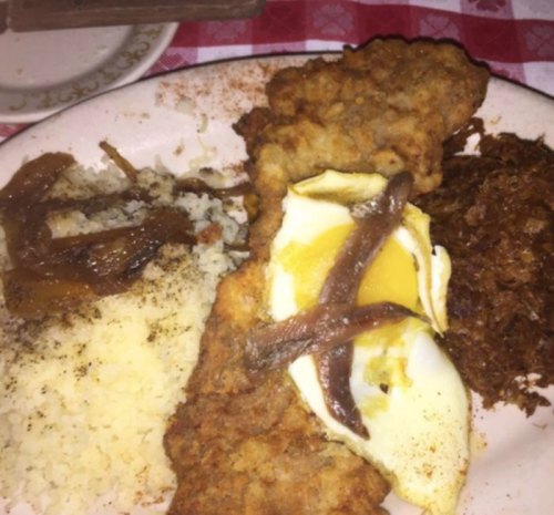 A plate of fried fish, rice, shredded meat, and a fried egg, garnished with sautéed onions on a checkered tablecloth.