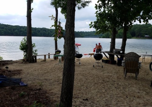 A sandy lakeside area with grills, chairs, and two people near the water, surrounded by trees and a cloudy sky.