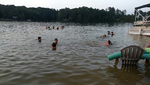 A group of people swimming in a lake, with trees and a boat in the background.