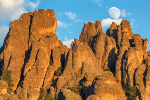 Rocky formations under a clear blue sky with a large moon rising above the peaks.