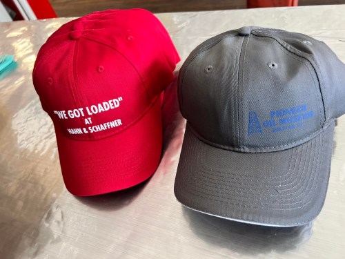 Two baseball caps: one red with "I'VE GOT LOADED" and the other gray with "PIONEER OILMUSEUM" embroidered.