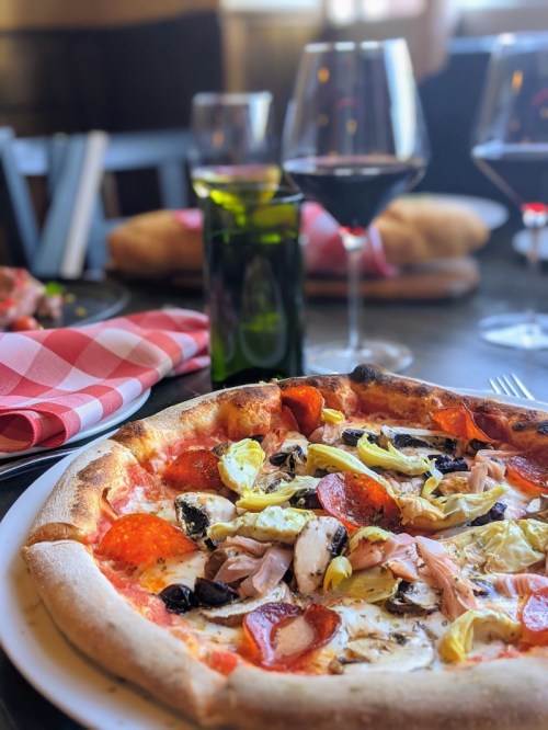 A close-up of a pizza topped with pepperoni, olives, and artichokes, with wine glasses and a checkered napkin in the background.