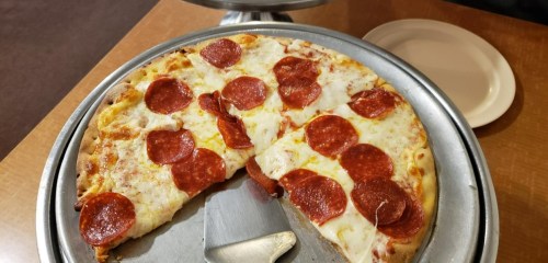 A pizza with pepperoni slices, partially sliced, on a metal tray with a plate in the background.