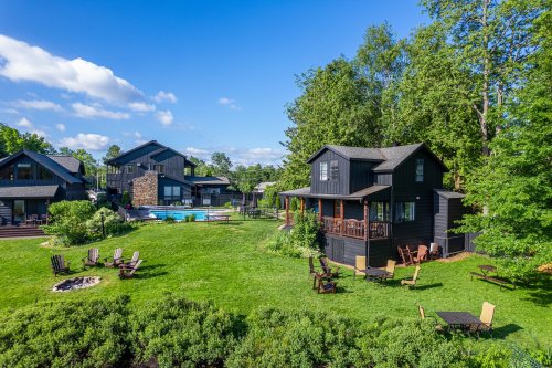 A scenic view of a black house surrounded by green grass, trees, and a pool, with outdoor seating and fire pit.