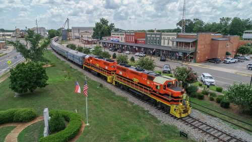 A colorful train passes through a small town with shops, trees, and flags under a cloudy sky.