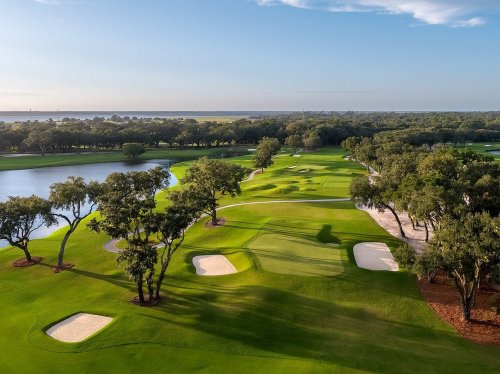 Aerial view of a lush golf course with sand traps, trees, and a lake under a clear blue sky.