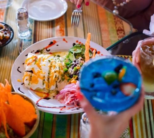 A colorful plate of enchiladas with salad and a vibrant blue drink in a lively restaurant setting.