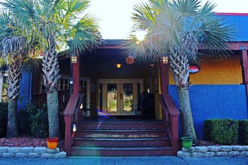 A colorful restaurant entrance with palm trees, steps, and an "Open" sign, bathed in warm sunlight.