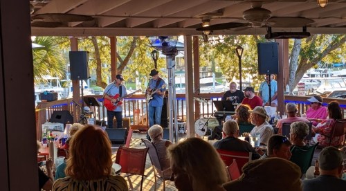 A lively outdoor music performance at a waterfront venue, with a band playing and an audience enjoying the show.
