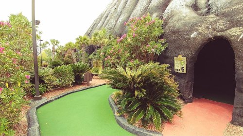 A mini-golf course with a green path, surrounded by tropical plants and a cave-like entrance.