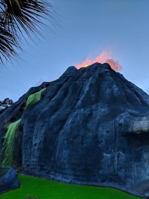 A volcanic structure with glowing orange at the top, surrounded by green water and palm leaves in the foreground.