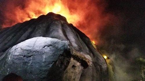 A volcanic eruption at night, with bright orange flames and smoke rising from a rocky mountain.