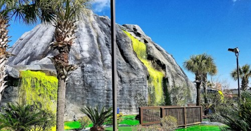 A large artificial rock formation with green waterfalls, surrounded by palm trees and a clear blue sky.