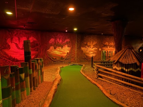 A dimly lit indoor mini-golf course with colorful murals, a winding path, and themed decorations.