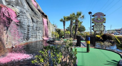 A vibrant mini-golf course featuring a waterfall, palm trees, and colorful landscaping under a clear blue sky.