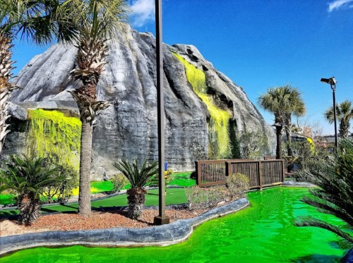 A mini-golf course featuring a rocky mountain, vibrant green water, palm trees, and a clear blue sky.