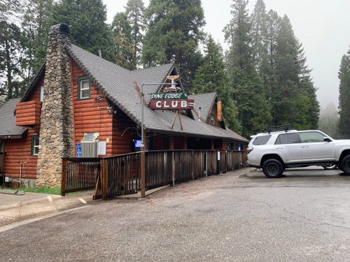 A rustic lodge with a sign reading "Pine Lodge Club," surrounded by tall trees in a foggy setting. A parked SUV is visible.