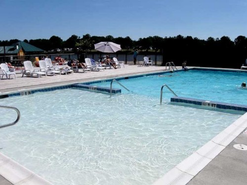 A sunny outdoor pool area with lounge chairs, umbrellas, and people swimming. Clear blue water and a bright sky.