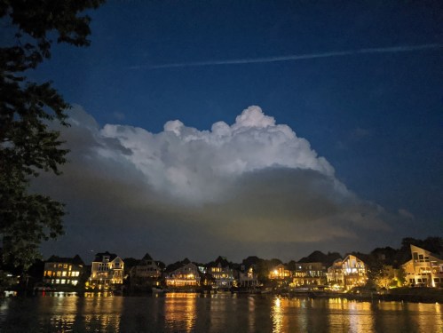 A serene night scene of a lake with illuminated houses and a large cloud in the sky, reflecting on the water.
