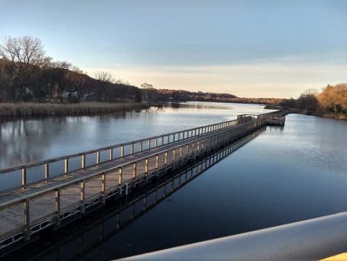 A serene river scene with a wooden walkway extending into the water, surrounded by trees and a clear sky.