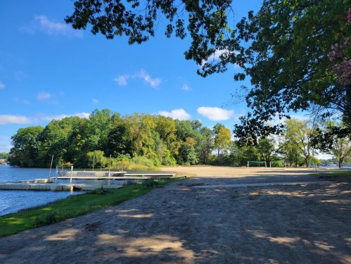 A serene lakeside view with a sandy area, trees in the background, and a clear blue sky.