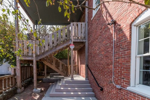 A wooden staircase leads to a second floor, beside a brick wall and a window, with greenery in the background.