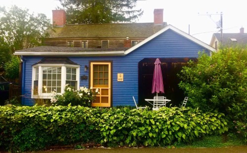 A charming blue house with a front garden, featuring a yellow door, white window, and a patio with a purple umbrella.