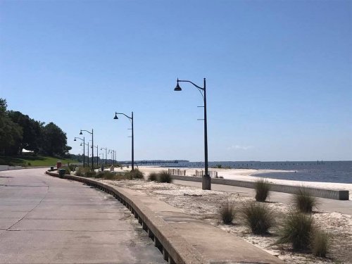 A sunny beachside walkway lined with lamp posts, leading to a calm sea and distant pier. Green trees in the background.