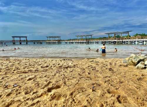 A sandy beach with people swimming and playing in the water, wooden docks in the background under a blue sky.