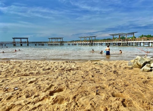 A sandy beach with people swimming and playing in the water, wooden docks in the background under a blue sky.