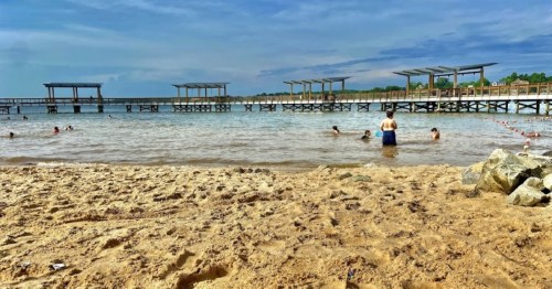 A sandy beach with people swimming and playing in the water, wooden piers in the background under a blue sky.