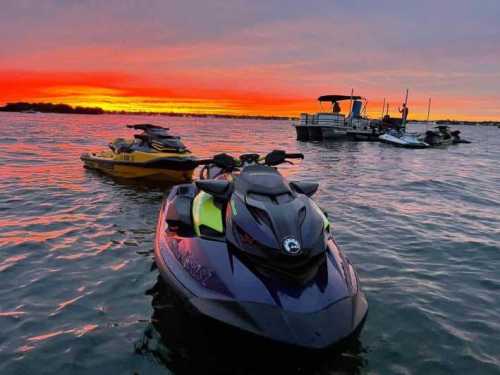 Two jet skis in the foreground on water, with a sunset sky and a boat in the background.